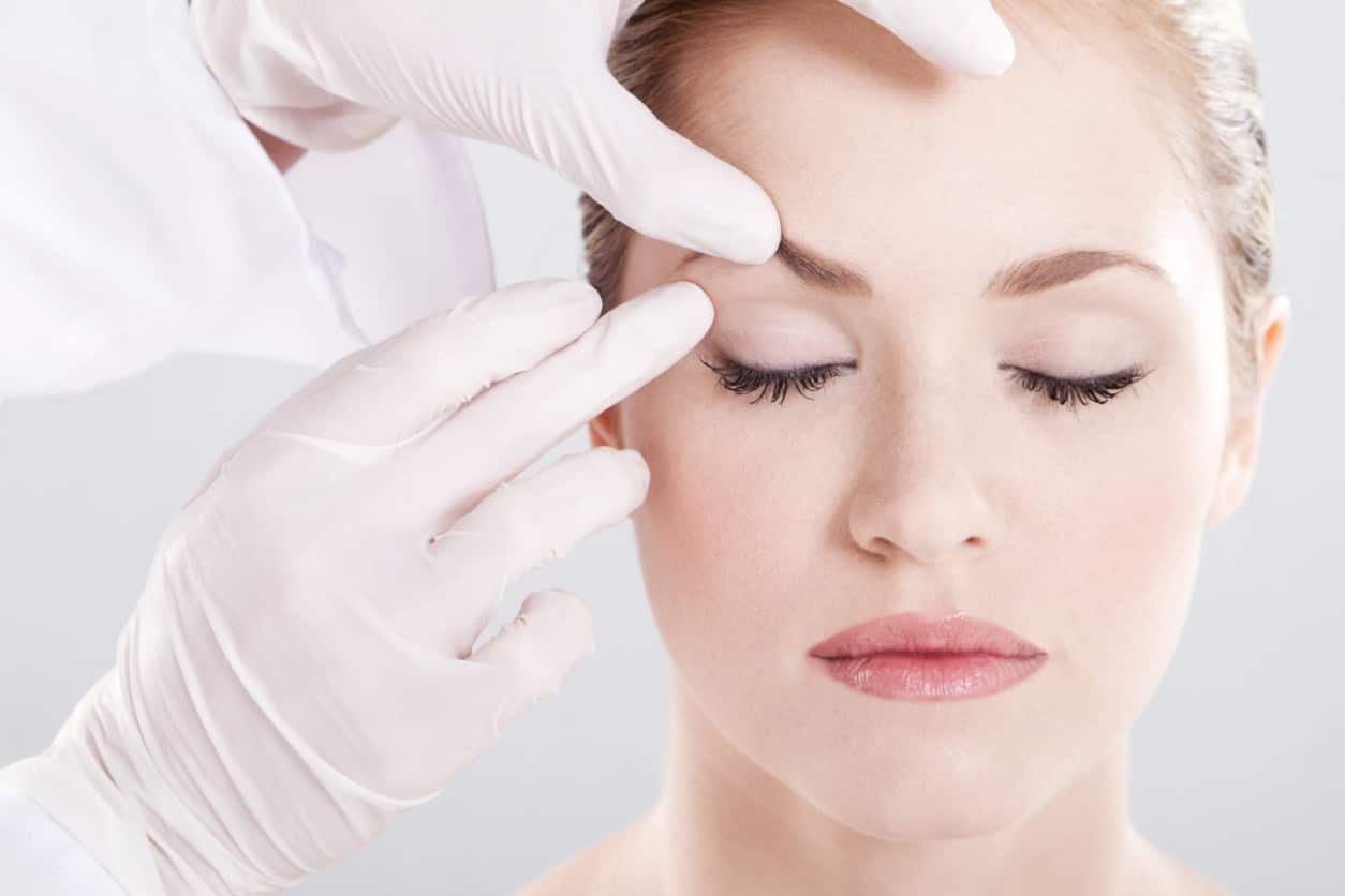 Headshot of woman with makeup on close her eyes and doctor in full scrubs examines her brow line, In doctor's office