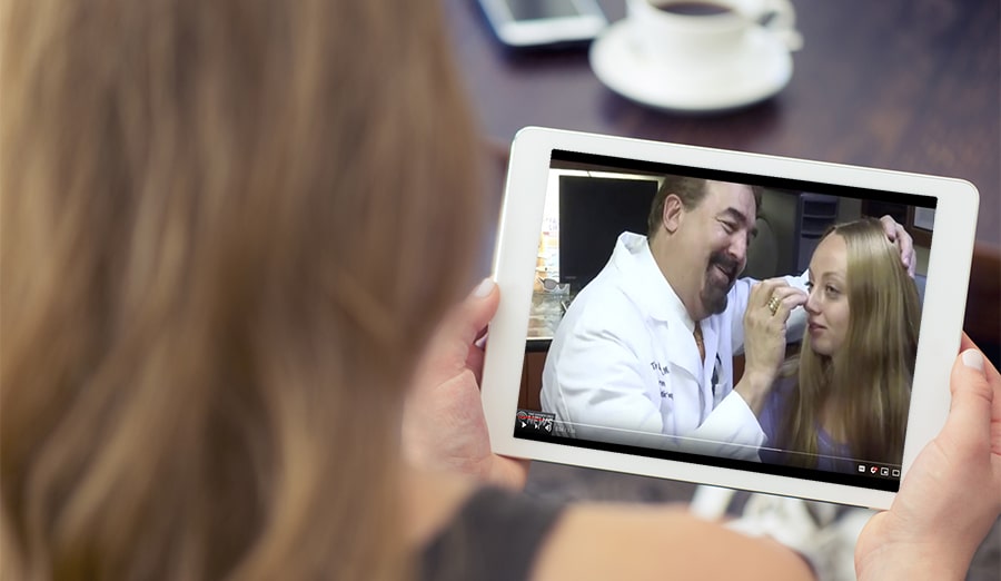 Woman looking at video. She is sitting in his living room on a sofa. Rear view over her shoulder so we can see the white screen. There are a coffee and mobile phone on the table